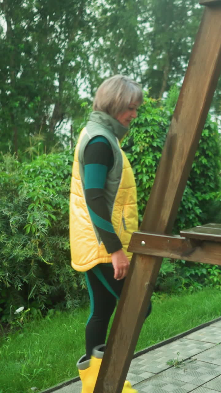 mujer en chaqueta amarilla y verde con botas amarillas camina hacia una bolsa de color menta descansando en un banco oscilante, el fondo presenta vegetación exuberante, jardín, árboles y un tambor de agua de plástico