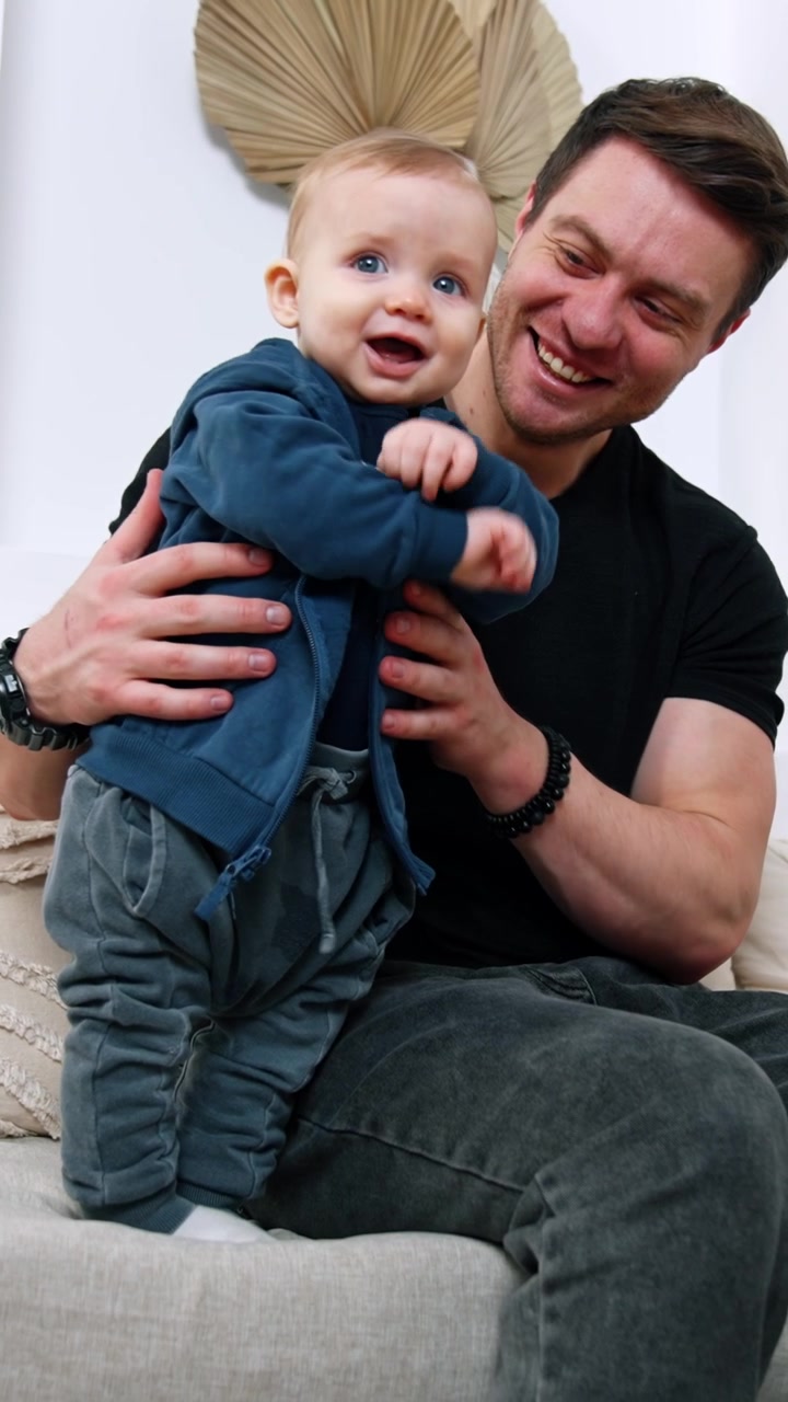 Happy Caucasian man sits on the sofa holding a baby boy. Smiling cute baby stands supported by father. Low angle view. Vertical video