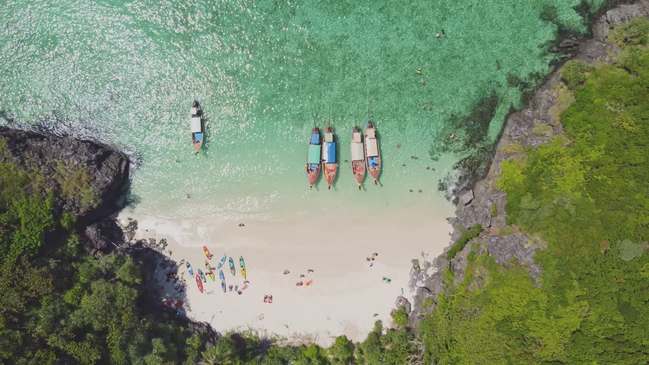 Aerial top-down view of a tropical beach on Koh Phi Phi with longtail boats, white sand, kayaks, and people enjoying the clear turquoise water in a calm, sheltered bay.