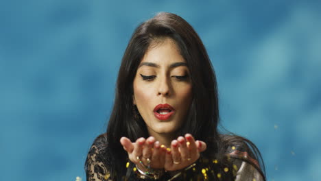 young stylish indian woman in traditional clothes with red lips blowing golden sparkles to camera