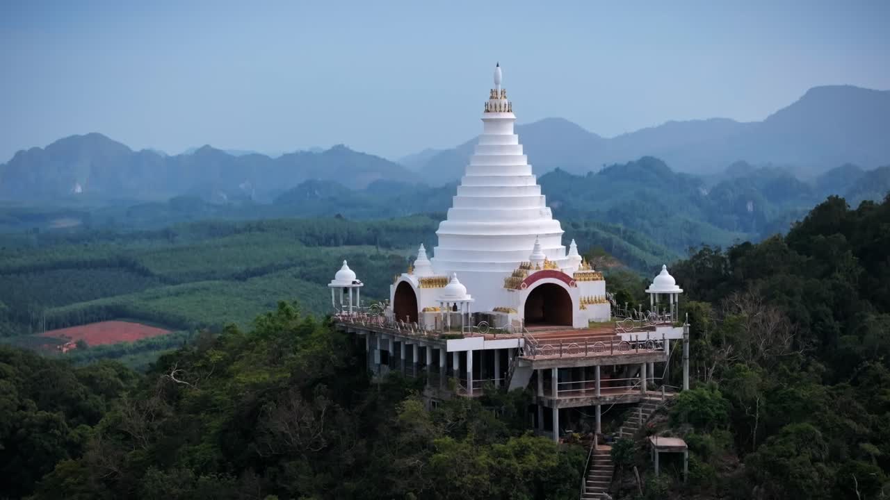 Drone footage of a white Buddhist temple atop a forested mountain in Surat Thani Province, Thailand. Midday light enhances the serene, spiritual landscape