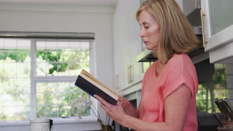 Mujer-Mayor-Caucásica-Parada-En-La-Cocina-De-Casa-Leyendo-Un-Libro