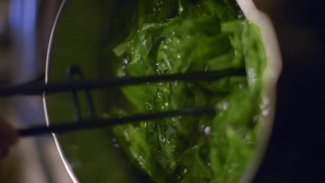 green vegetables in pot of boiling water stirred with tongs, filmed as vertical closeup slow motion handheld style shot