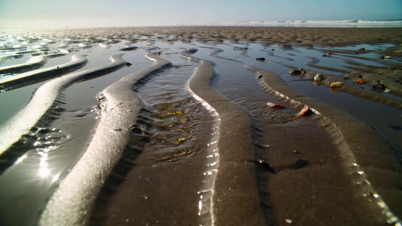 Retreating tide pulling water on low-tide beach, revealing wet rippled sand ridges, foam lines