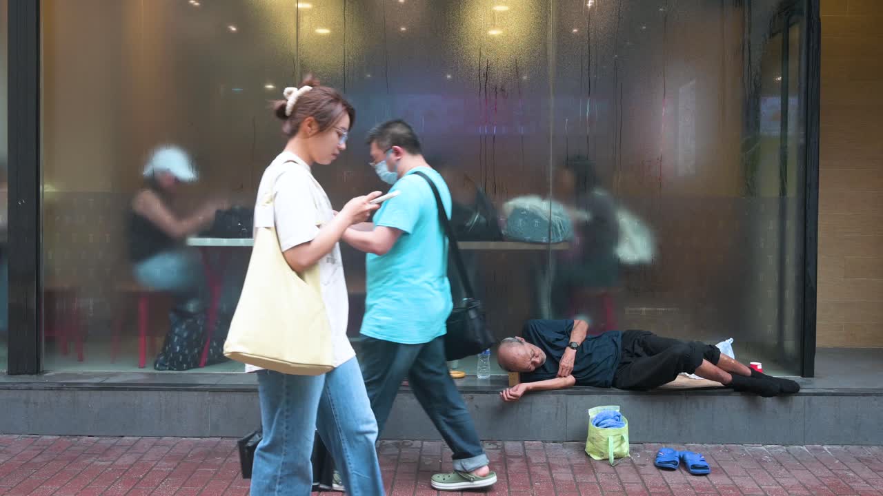 A homeless elderly man rests outside a restaurant, with diners visible through the window, as pedestrians move by in the bustling streets of Hong Kong.