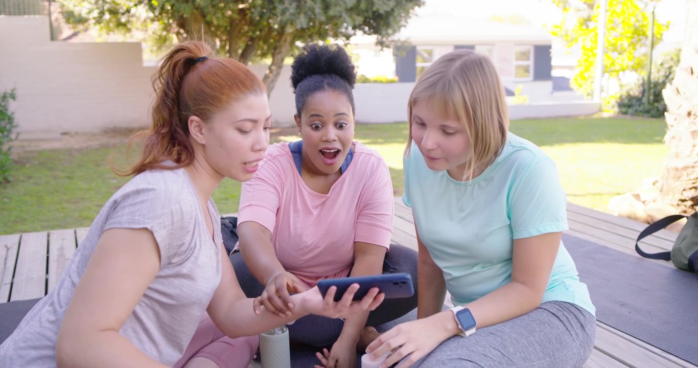 Three female friends in leggings taking selfies, reviewing photos on deck with phone and yoga mats