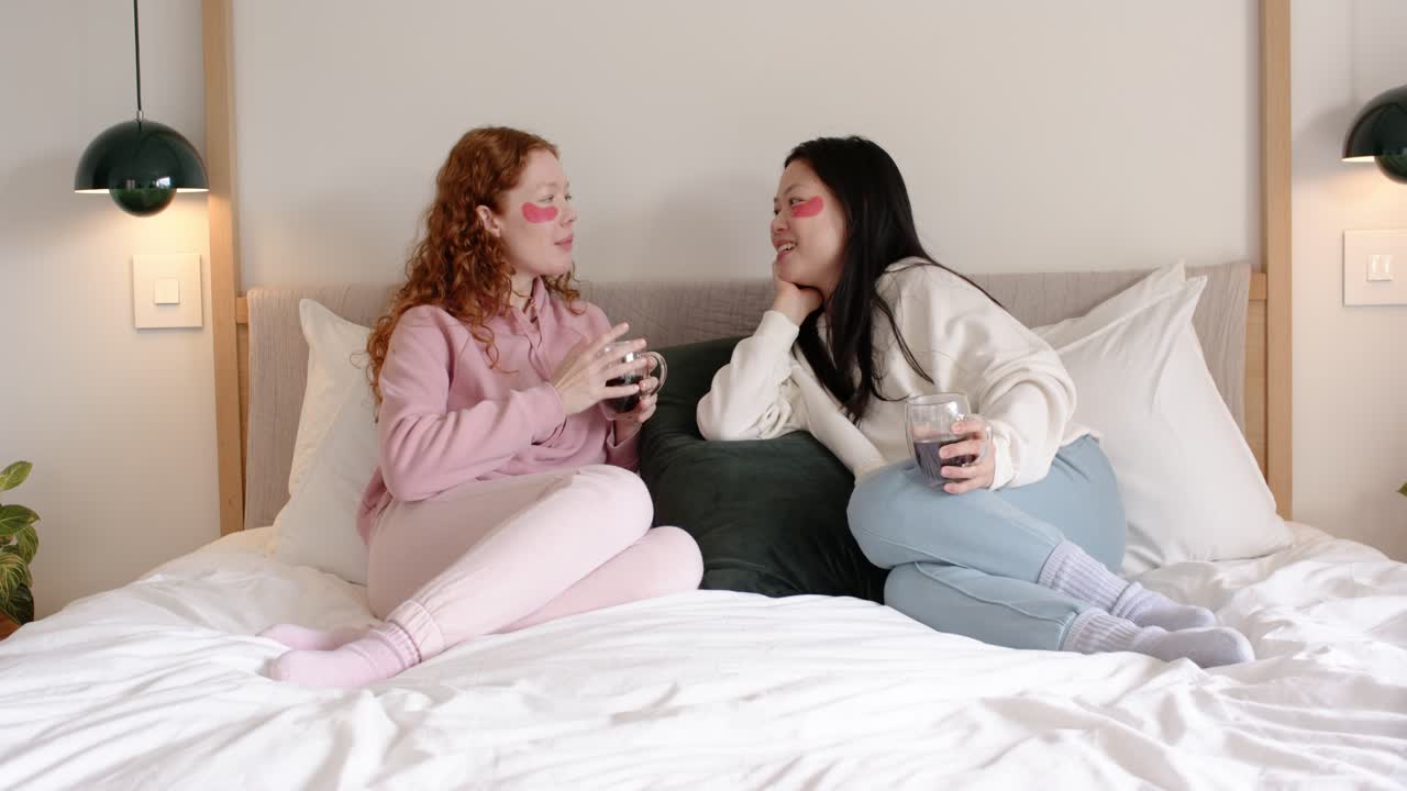 Diverse female pair starting intimate chat, touching hands and sipping on bed with undereye patches