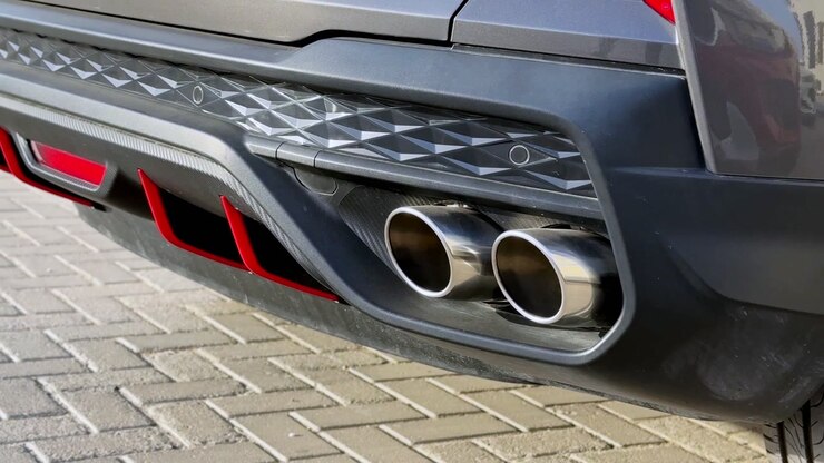 Primo piano di doppi tubi di scarico cromati sul paraurti posteriore di un'auto sportiva parcheggiata su terreno pavimentato. Scatto dettagliato che mostra il design automobilistico e gli accenti del diffusore posteriore durante il giorno