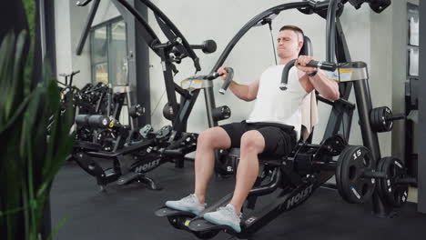 Young man working out on weight machine doing chest press with energetic facial expression and flexing muscles, flower vase visible in corner of modern gym with sleek equipment under bright lighting