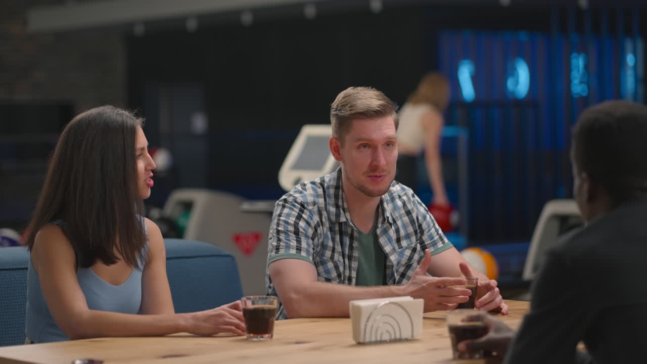 A multi-ethnic group of friends sits at a table together in a bowling alley and talks
