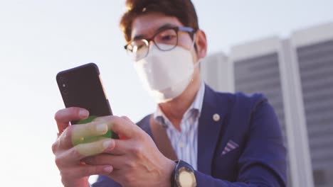 Asiatischer-Mann-Mit-Gesichtsmaske-Und-Smartphone,-Während-Er-Auf-Der-Treppe-Im-Firmenpark-Sitzt