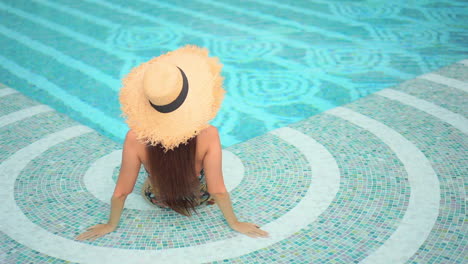 Sitting-in-the-shallow-end-of-the-pool-with-her-back-to-the-camera,-a-young-woman-turns-her-head-to-survey-the-water