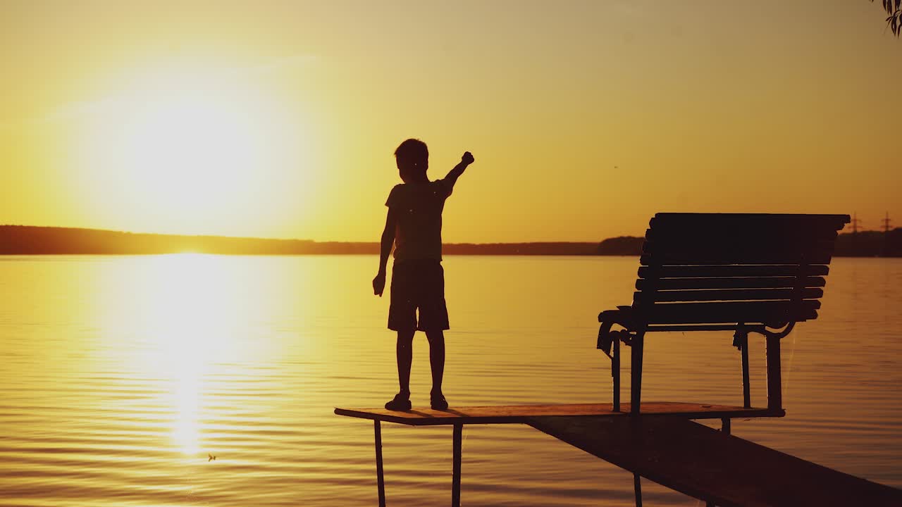 A little boy is standing on the masonry near the bench and doing physical exercises. The child is raising hands up alternately on the background of the river at sunset.