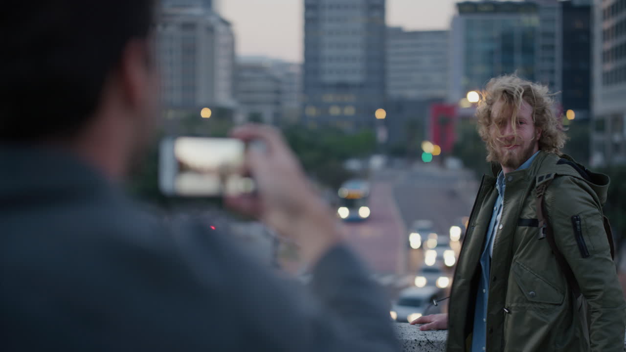 초상화 행복한 젊은 남자가 사진을 찍기 위해 포즈를 취하고 팔을 들어 올려 뛰어 성공적인 여행 휴가 라이프 스타일을 축하하고 웃는 친구가 스마트 폰을 사용하여 도시에서 사진을 찍습니다.