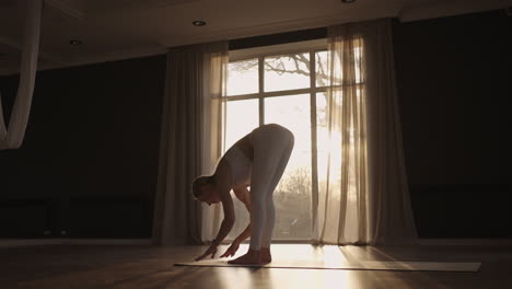 slow motion: young woman is doing yoga in a white room filled with light the girl performs yoga stands and elements near the large window.