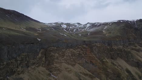 Aerial-panoramic-landscape-view-of-mountain-rock-formations-and-steep-cliffs,-with-melting-snow,-on-a-moody-evening-in-Iceland