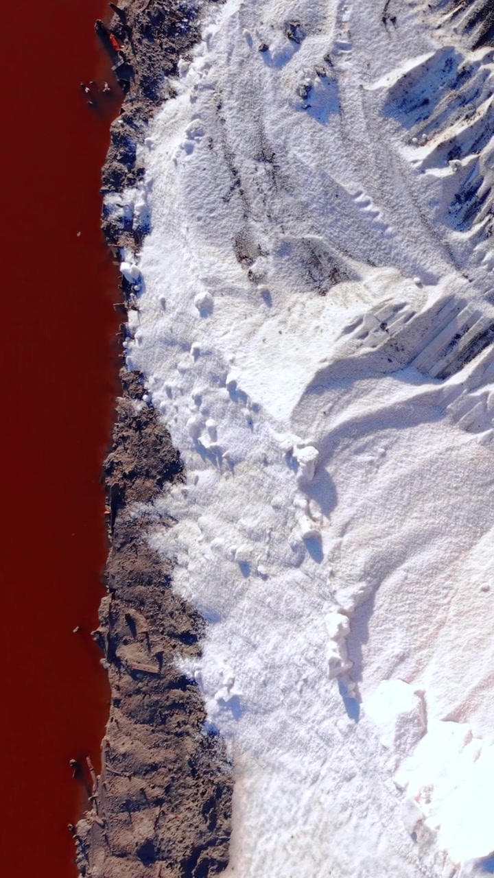 Vista aerea ravvicinata di uno stagno rosso e del bordo di sale. Acqua salmastra rosso intenso incontra un mucchio di sale cristallizzato bianco sulla riva. Video verticale.