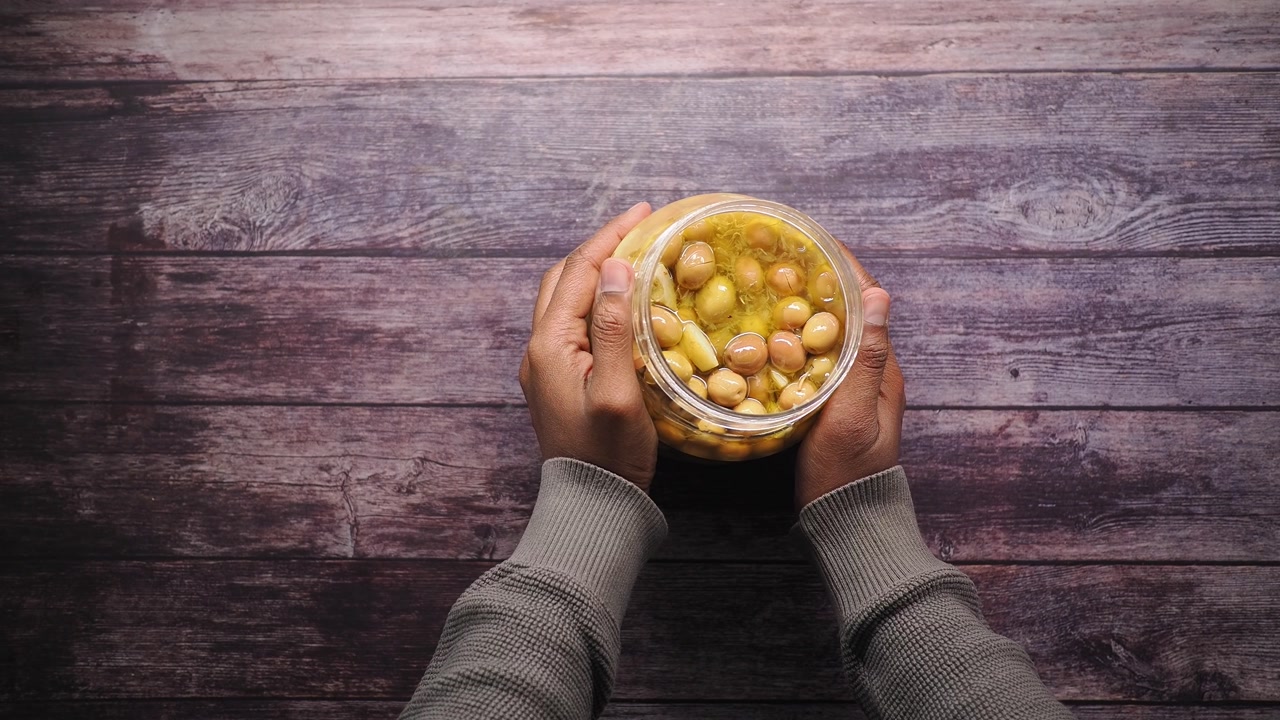 Hands Holding a Jar of Pickled Chickpeas and Garlic on a Wooden Table