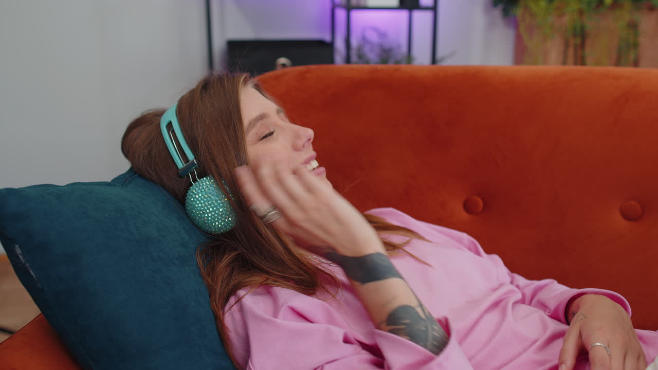무선 헤드폰을 입은 행복한 젊은 여성이 집에서 소파에 누워서 좋아하는 음악을 듣고 휴식을 취하고 있습니다.