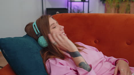happy young woman in wireless headphones relaxing lying on sofa at home listening favorite music
