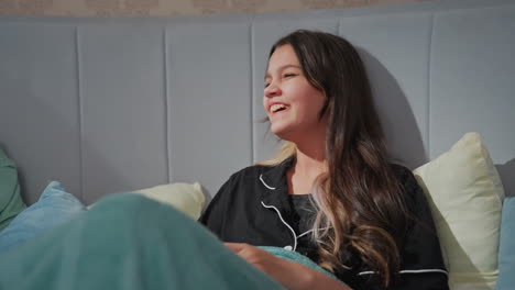 close up of joyful young girl with long dark hair laughing with mouth partly covered while eating popcorn and chatting with friend, cozy bedroom setting with soft pillows and relaxed atmosphere