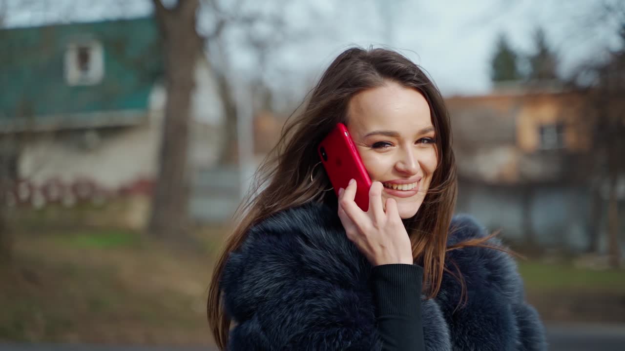 Portrait of a smiling girl with a phone. Beautiful young woman dressed in fur coat seductively poses for camera and talks the phone in the city park. Motion camera around.