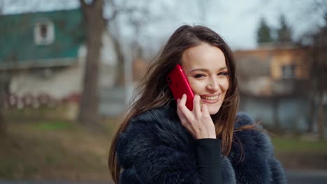portrait of a smiling girl with a phone. beautiful young woman dressed in fur coat seductively poses for camera and talks the phone in the city park. motion camera around.