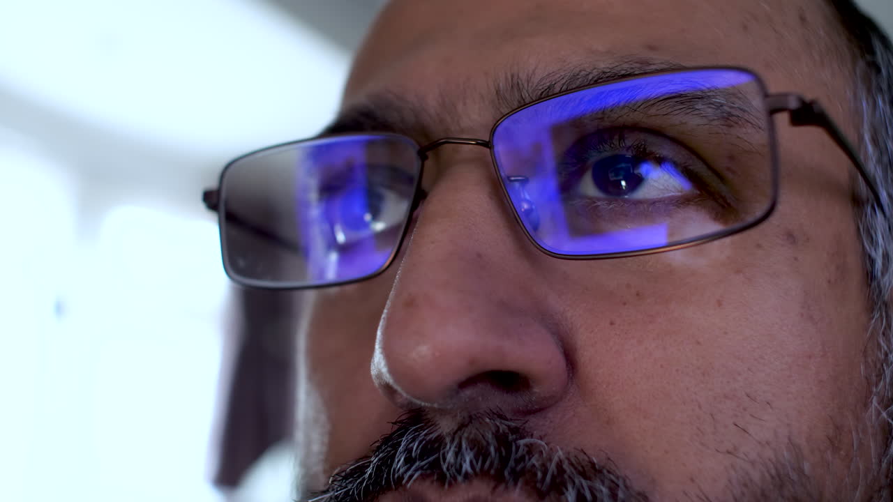 Close up of a South Asian man wearing blue light glasses with the reflection of a computer screen visible in the lenses