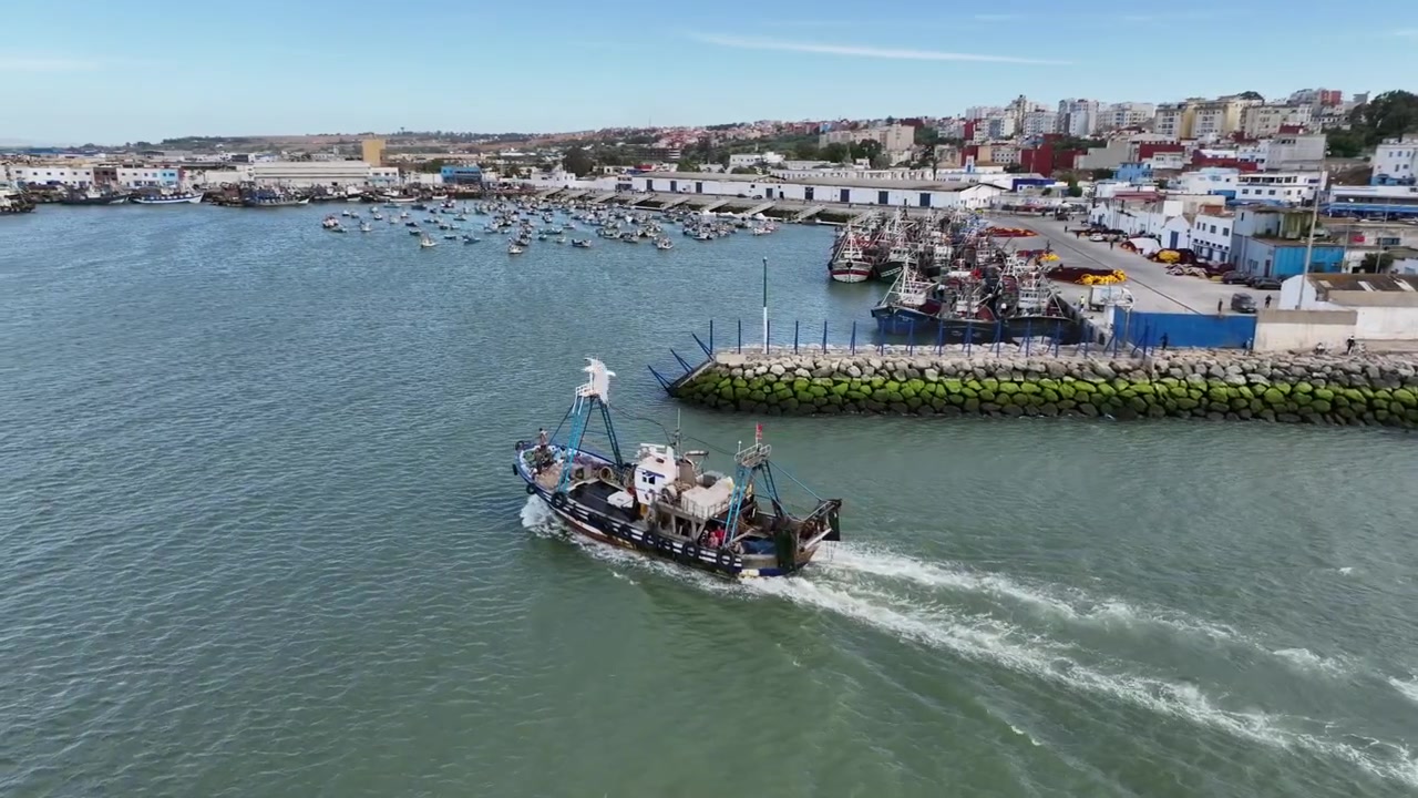 Vista aerea in movimento di un peschereccio in attività che si muove attraverso un porto atlantico in Marocco, lasciando una scia con moli da pesca, flotta e città costiera sullo sfondo.