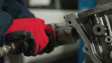 engineer wearing red gloves operates pneumatic tool on engine block in industrial workshop, close-up captures mechanical repair process, and metal components with blurred white vehicle in background