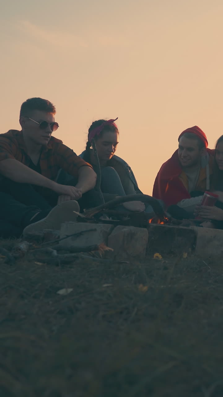 cheerful young hikers group rests at camping with burning bonfire and tent on river bank at autumn evening twilight