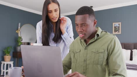 Video-of-diverse-couple-in-bedroom-drinking-coffee-and-looking-at-laptop-screen-together