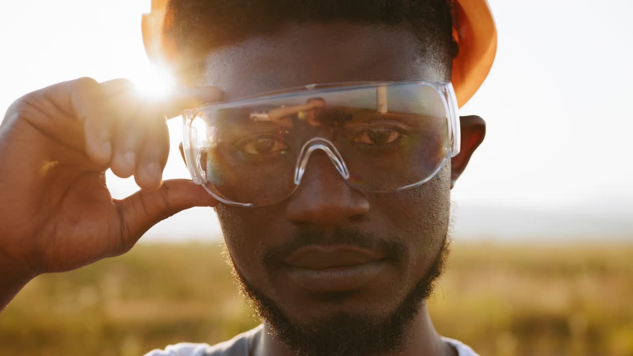 hombre afroamericano de pie en el campo con enormes molinos de viento durante la puesta del sol de verano. técnico profesional con mono gris, casco naranja y gafas de seguridad. concepto de energía alternativa