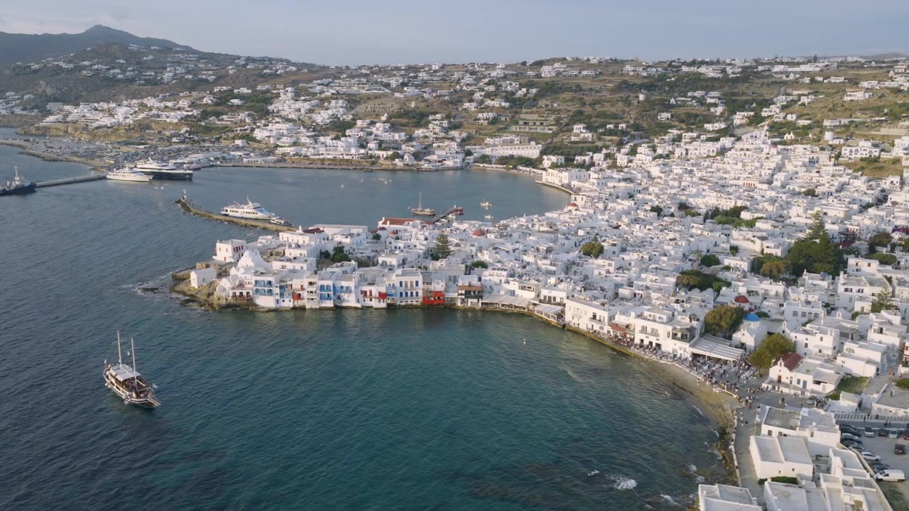 Ampia veduta aerea della città di Mykonos e del suo vecchio porto, con barche all'ancora, fitti edifici imbiancati a calce e colline che si ergono sullo sfondo.