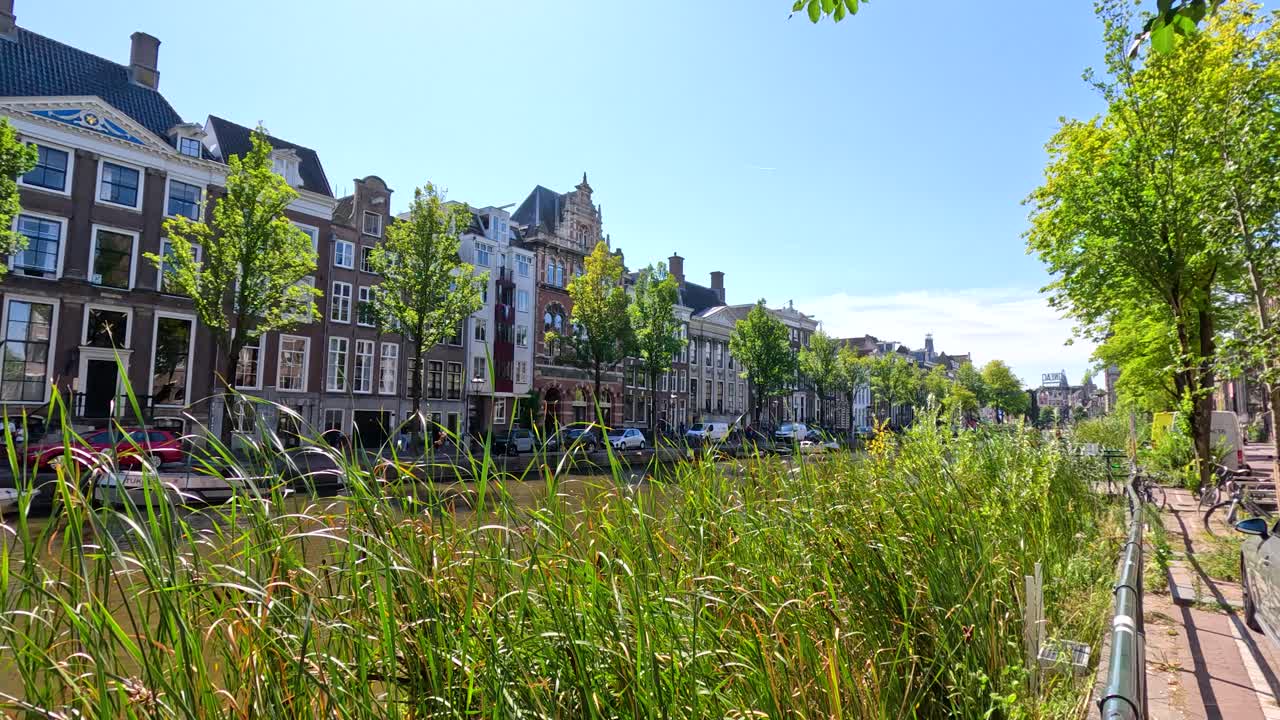 Camera moves along canal, showing historic Dutch houses, greenery, reflections, and bright daylight
