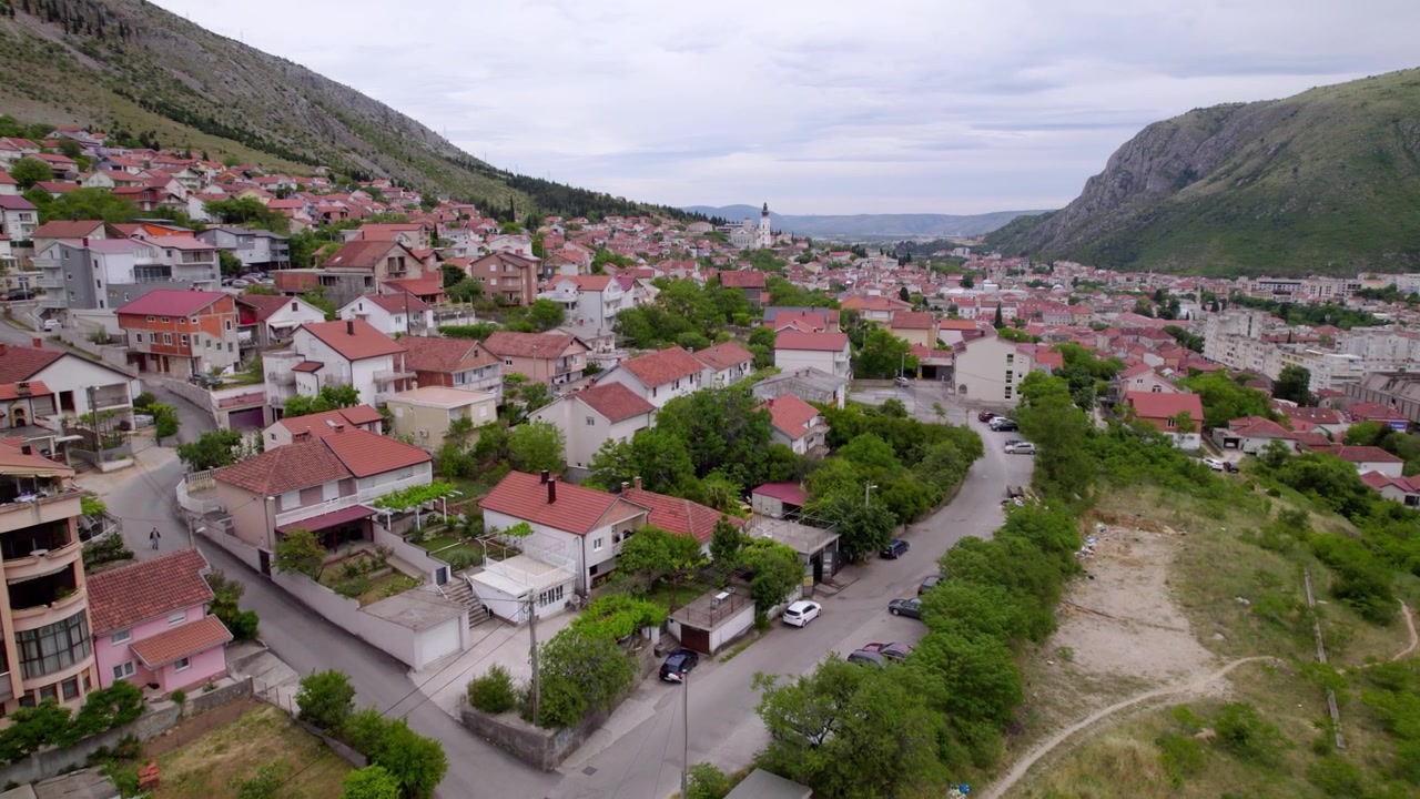 Ilmakuva Mostarin kaupunginosista vuorenrinteiden välissä, mutkittelevine paikallisteineen ja oransseine, hillittyine kattoineen, Bosnia ja Hertsegovinassa.