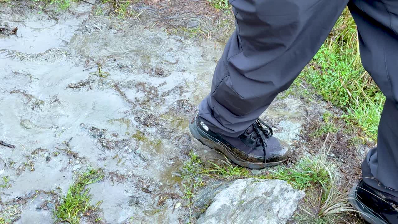 Person in outdoor gear steps carefully along wet, muddy path with overcast natural lighting
