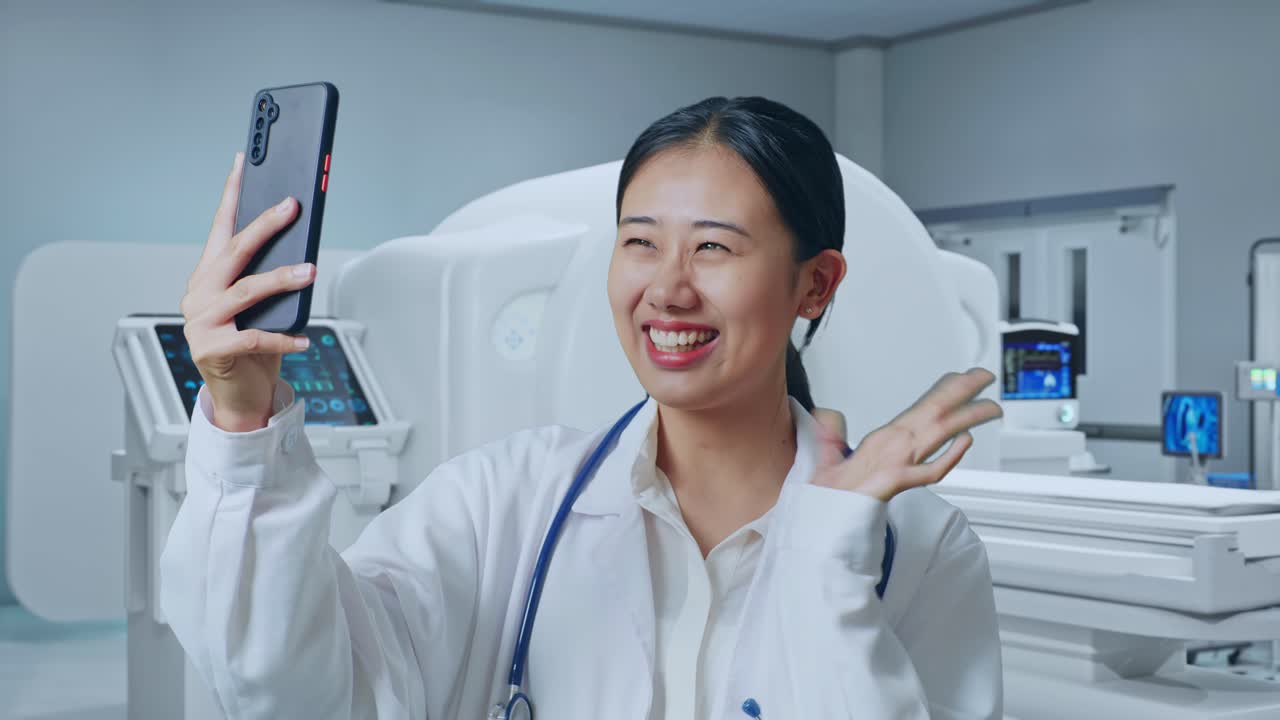 Close Up Of Asian Woman Doctor With Stethoscope Waving Hand And Having A Video Call On Smartphone While Standing With Mri Machine In The Hospital