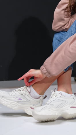 Cropped image of a girl sitting on a black step. Model in jeans, sweatshirt and white sneakers in studio. Vertical video