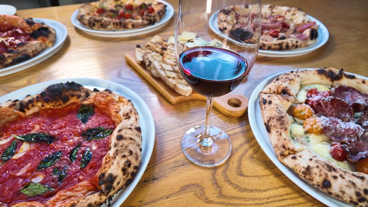 Ripresa panoramica dall'alto di pizze italiane, vino rosso e antipasti su un tavolo di legno.