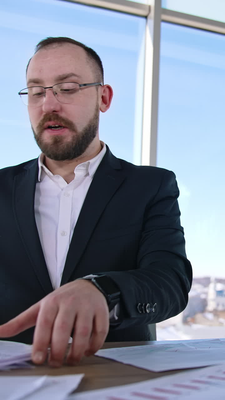 Successful entrepreneur speaking in office. Businessman in glasses and elegant suit sitting at the table in his workplace and talking seriously to someone. Vertical video