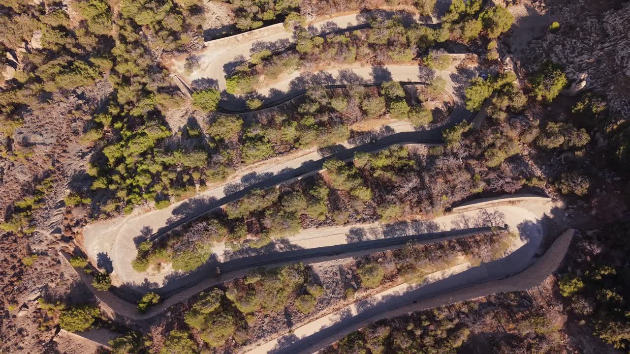 Top-down drone view of a zigzag road in Santorini with a 4-wheel bike moving along the winding path, showcasing cliffs, caldera, and the stunning island landscape