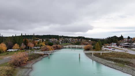 Aerial view of Lake Tekapo's turquoise waters and bridge, surrounded by autumn foliage under overcast skies. Captures serene landscape and infrastructure