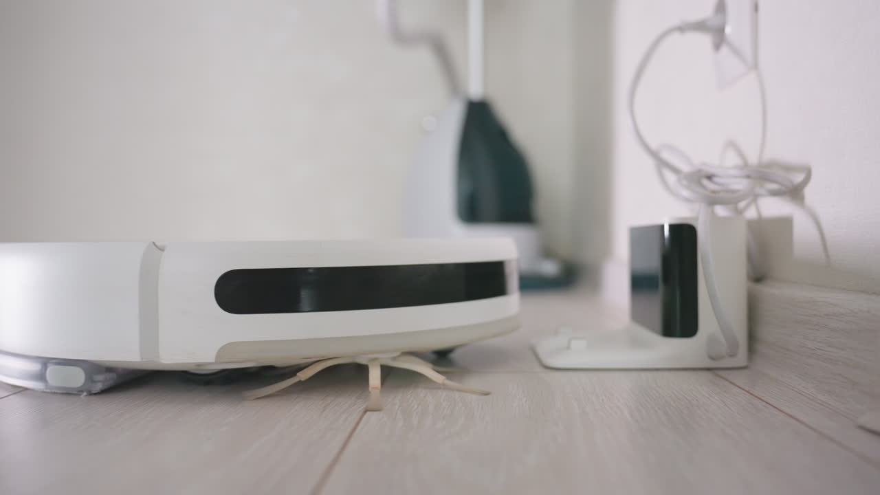 Hand presses button to activate robot vacuum cleaner as it moves away from charging dock on wooden floor, showing modern automated cleaning technology in home environment
