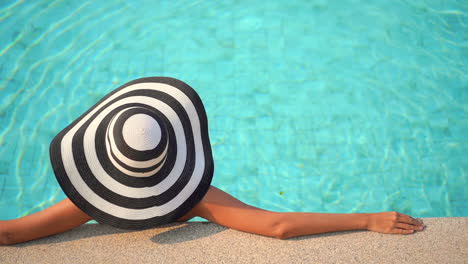 a woman with her back to the camera and wearing a huge black and white floppy sun hat relaxes in a swimming pool