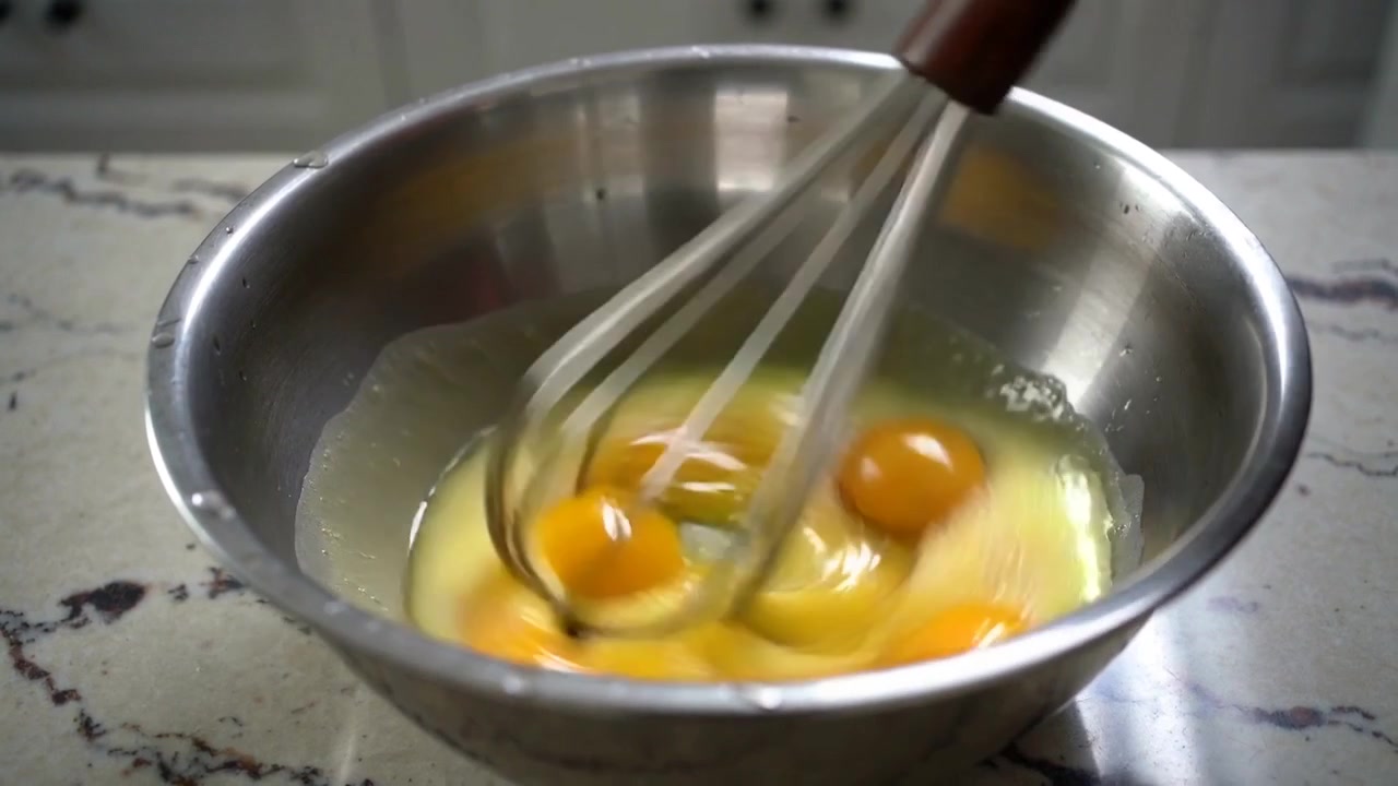 Pengocok kawat masuk dari kanan atas, mengocok telur yang sudah dipecahkan dalam mangkuk baja di meja dapur di rumah untuk persiapan.