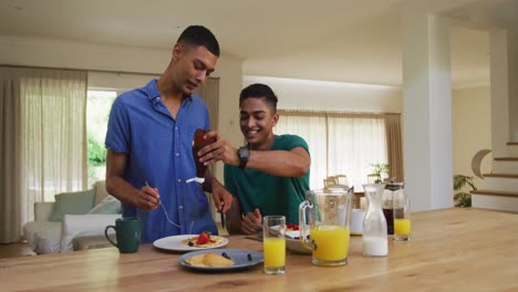 Smiling-mixed-race-gay-male-couple-having-pancakes-for-breakfast-in-kitchen