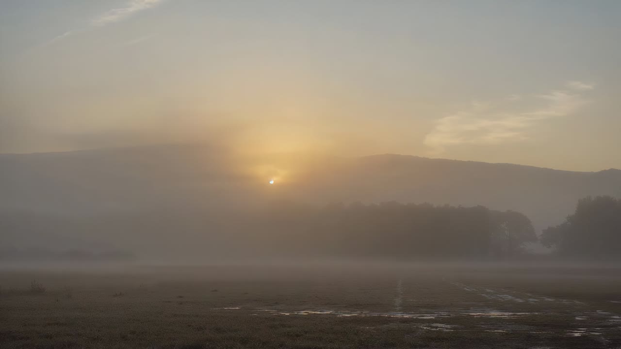 Rising sun breaking low fog bank over hill at dawn, illuminating waterlogged field and puddles