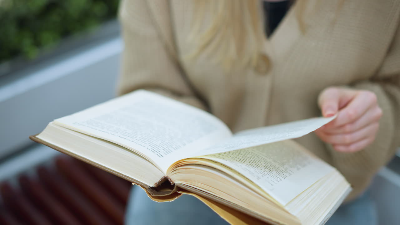 primer plano de una mujer con un suéter tejido de oro volviendo a la siguiente página del libro, mano en la página, flor decorativa verde ligeramente borrosa en el fondo