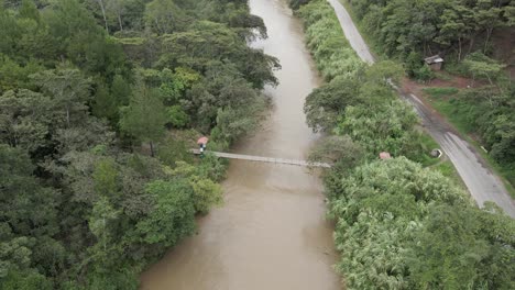 Sobrevuelo-Aéreo-De-Carretera-Estrecha-Y-Puente-Peatonal-Sobre-El-Río,-Selva-Peruana